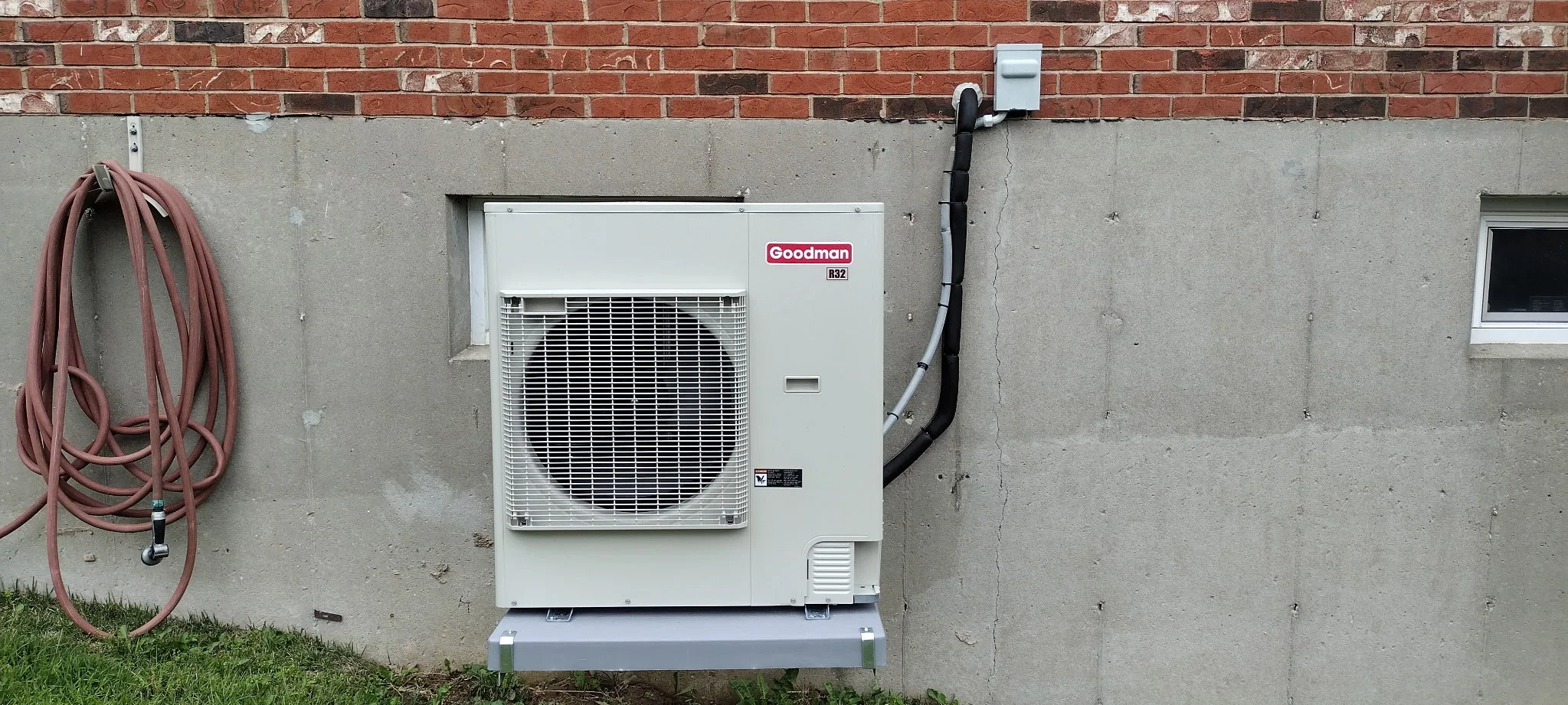 Goodman R32 heat pump unit installed on concrete pad with black refrigerant lines and disconnect switch